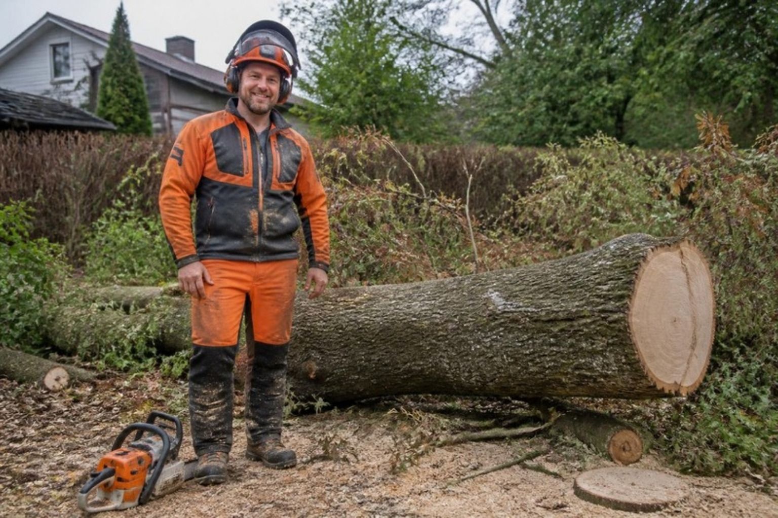 Smilende træfælder i orange skovtøj og hjelm står ved en fældet træstamme i en have